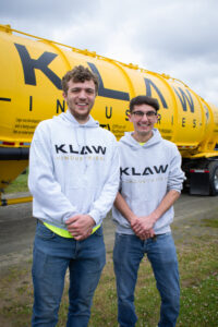 Two men standing in front of large vehicle labeled: KLAW Industries