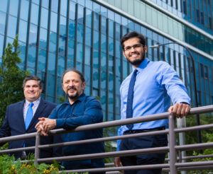 Three men smiling, leaning over a railing outside of an office building.